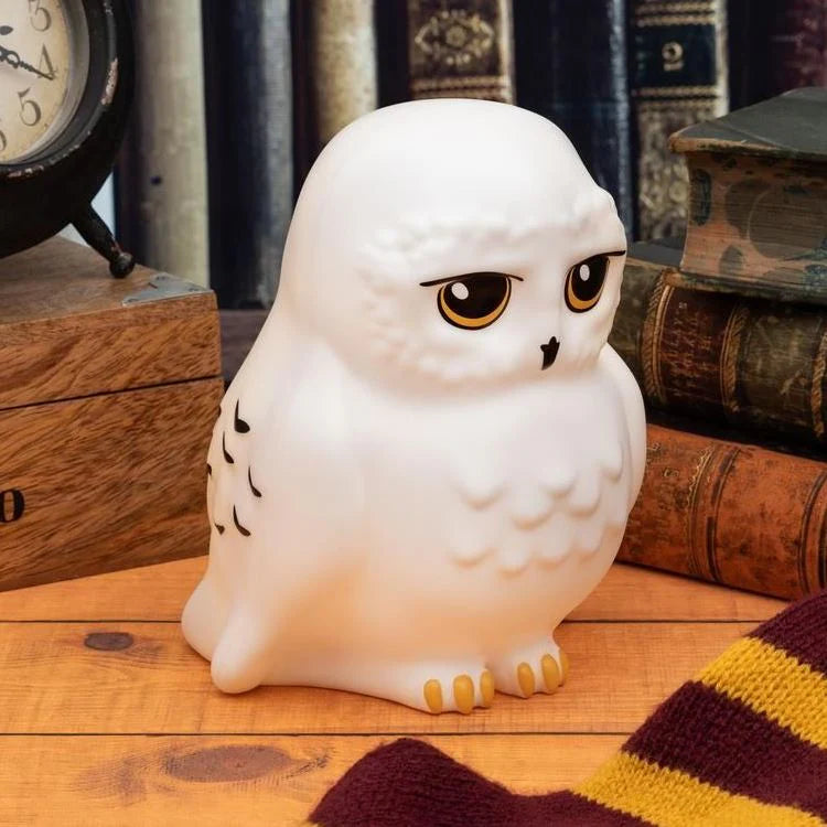 The Harry Potter Hedwig Light by Paladone features a white owl with yellow eyes and black markings, perched on a wooden surface amid vintage books, a scarf, and clock—perfect for any Harry Potter fan’s collection.