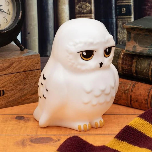 The Harry Potter Hedwig Light by Paladone features a white owl with yellow eyes and black markings, perched on a wooden surface amid vintage books, a scarf, and clock—perfect for any Harry Potter fan’s collection.