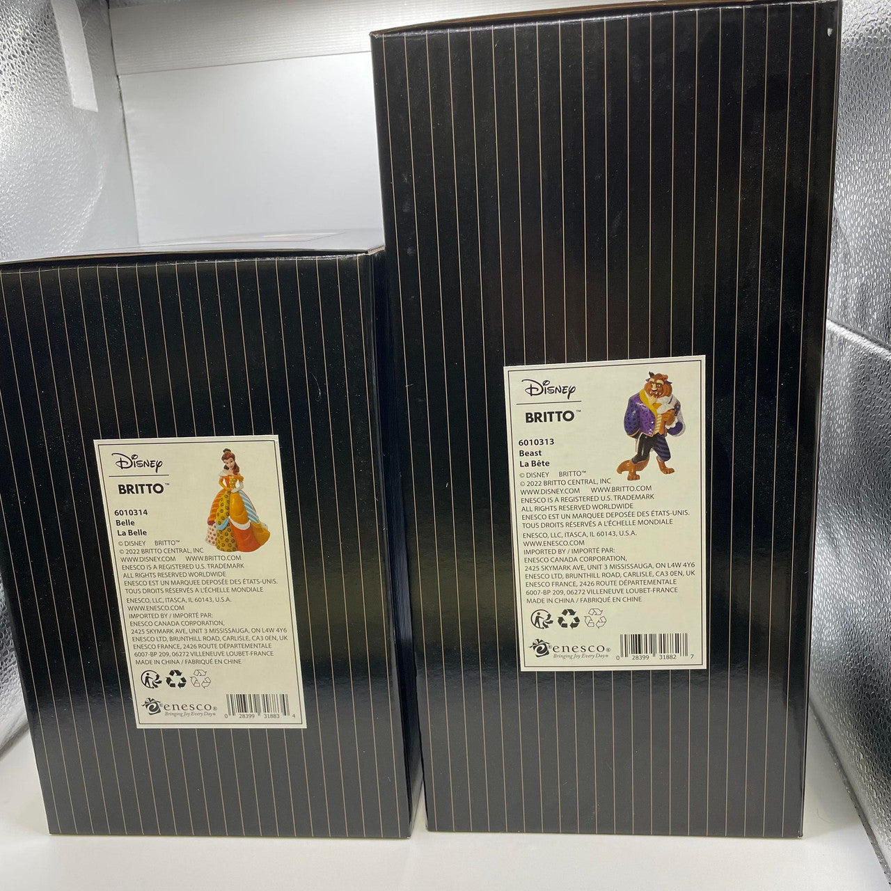 Two black, vertically striped Disney Britto boxes, each labeled with Belle and Beast figurines from the Disney Britto Belle and Beast Double Pack, stand upright in a lighted display area.