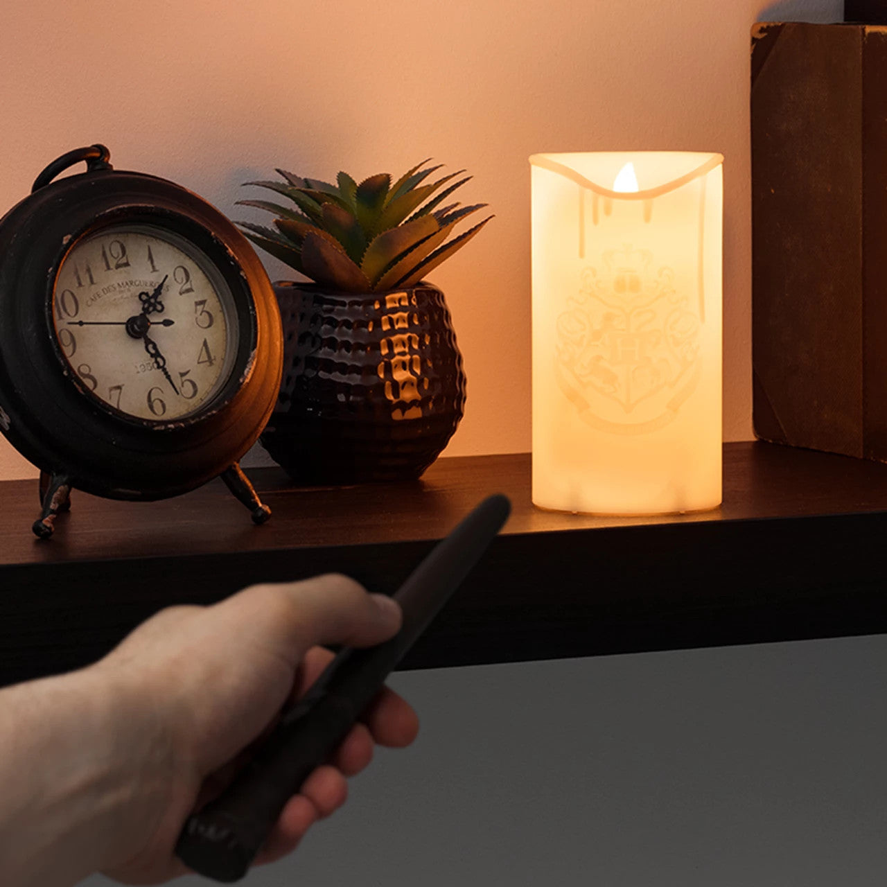 A hand aims the wand remote at the lit Harry Potter Candle With Wand Remote Control Light By Paladone, displayed on a shelf with a vintage clock, Hogwarts crest, potted plant, and book. Brand: Paladone.