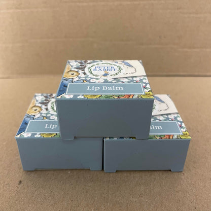 Three Beatrix Potter Peter Rabbit Lip Balm Triple Packs are stacked in a pyramid against a plain cardboard background. Each Beatrix Potter box shows charming illustrations and a “Lip Balm” label on the front.