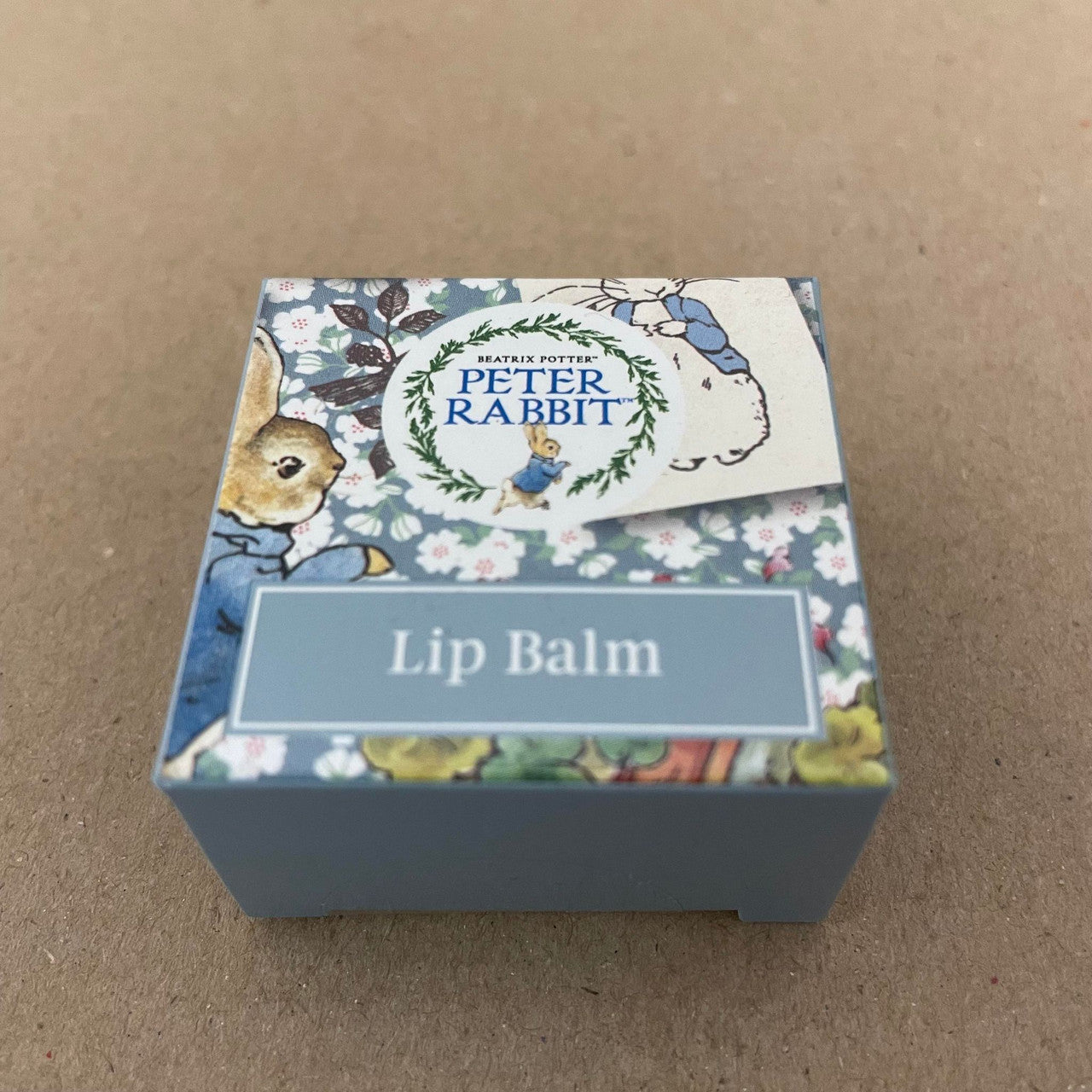 A small square Beatrix Potter Peter Rabbit Lip Balm Triple Pack gift box, decorated with charming rabbits, floral patterns, and the words Lip Balm and Beatrix Potter PETER RABBIT on the lid, sits on a plain brown surface.