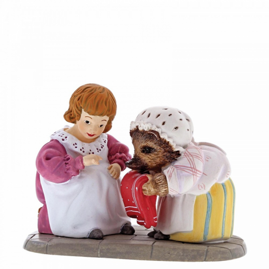 The Beatrix Potter Mrs. Tiggy Winkle and Lucie Figurine features Lucie in a pink dress kneeling beside Mrs. Tiggy Winkle, both gazing at a red suitcase on a gray oval base—a charming collectible from the Beatrix Potter brand.