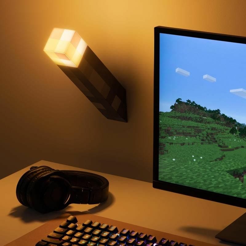 The Paladone Minecraft Torch Light, which mounts on the wall or stands on a table, glows brightly above a Minecraft game on a monitor, with a keyboard and headphones organized neatly on the desk below.