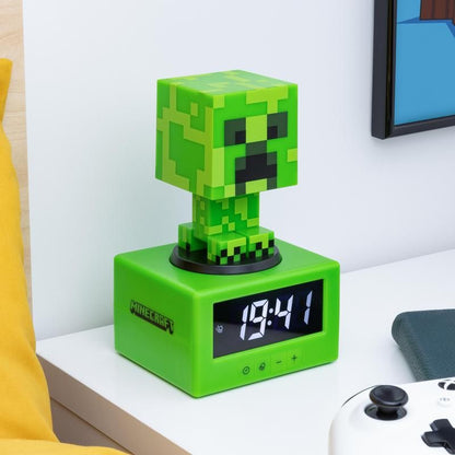 The Minecraft Creeper Icon Alarm Clock by Paladone shows 13:41 on a white bedside table in a gamers bedroom, next to a white game controller and a book.