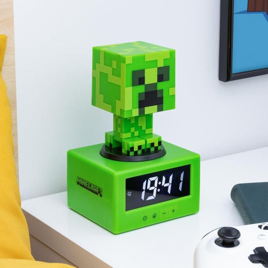 The Minecraft Creeper Icon Alarm Clock by Paladone shows 13:41 on a white bedside table in a gamers bedroom, next to a white game controller and a book.