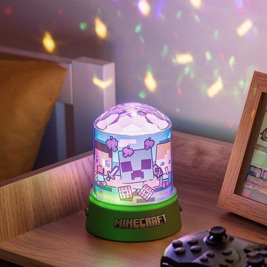 The Minecraft Party Projection Light by Paladone sits on a wooden bedside table, casting colorful pixelated characters and blocks on the wall. The green base is labeled Minecraft, and a game controller rests nearby.