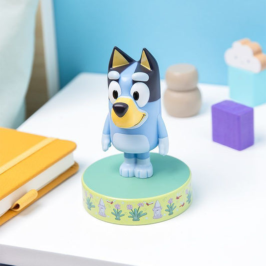 The Bluey Icon Light by Paladone, shaped like a blue heeler puppy, stands on a decorated base atop a white table with a yellow notebook, geometric blocks, and a cloud ornament in a bright room.