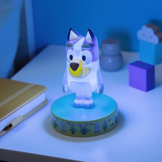 The Bluey Icon Light By Paladone, shaped like a blue heeler puppy, stands on a decorated base on a white bedside table beside a notebook and colorful blocks in a softly lit room.