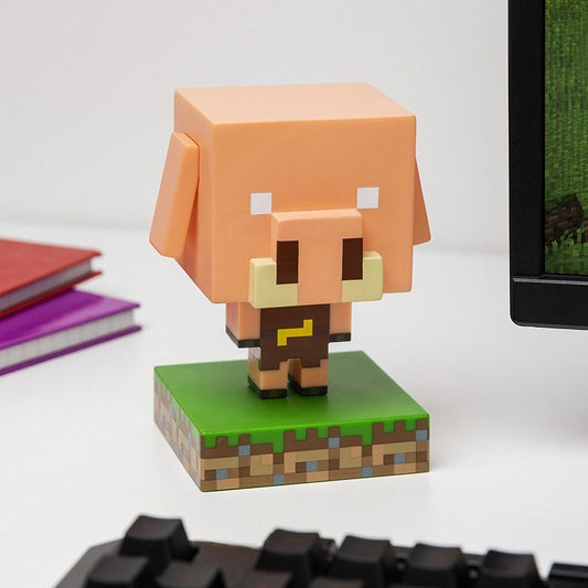 The Minecraft Piglin Icon Light by Paladone, a blocky pig-like collectible, stands on a green pixelated base atop a white desk near a computer monitor with colorful notebooks in the background.