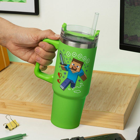 A hand holds the Paladone Minecraft Insulated Cup and Straw Steve & Creeper, a large green tumbler with handle and graphic icons. It has a clear multi-way lid with straw and sits on a wooden desk near office supplies and a monitor.