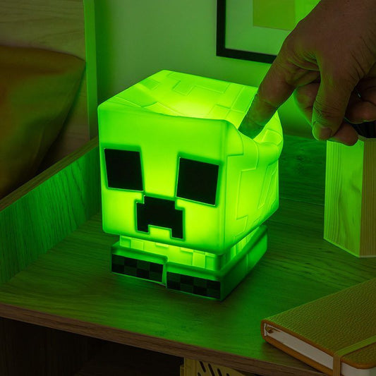 The Minecraft Creeper Squishy Glo Silicone Rechargeable Light by Paladone sits on a wooden nightstand as a hand presses down on its glowing green head.