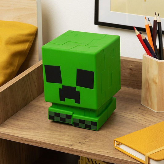 The Minecraft Creeper Squishy Glo Silicone Rechargeable Light by Paladone sits on a wooden desk beside a yellow pillow, a cup of colored pencils, and a closed yellow notebook.