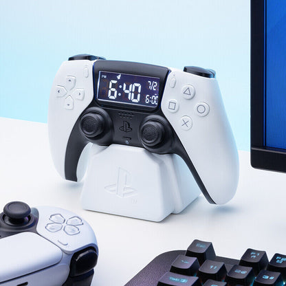 A white Playstation 5 Controller Alarm Clock by Paladone sits on its stand, flanked by another controller and part of a gaming keyboard and monitor on a tidy desk.
