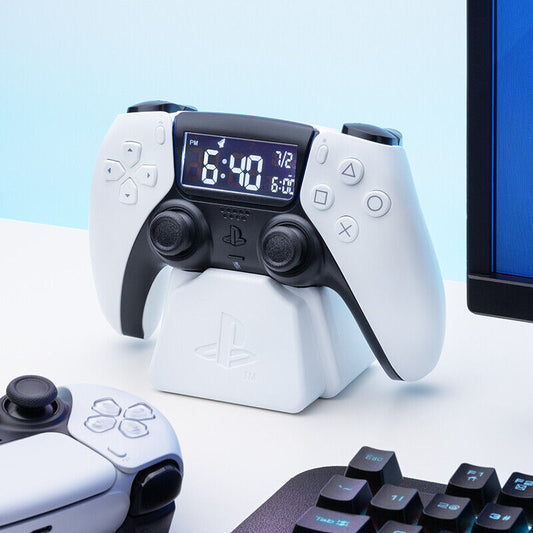 A white Playstation 5 Controller Alarm Clock by Paladone sits on its stand, flanked by another controller and part of a gaming keyboard and monitor on a tidy desk.