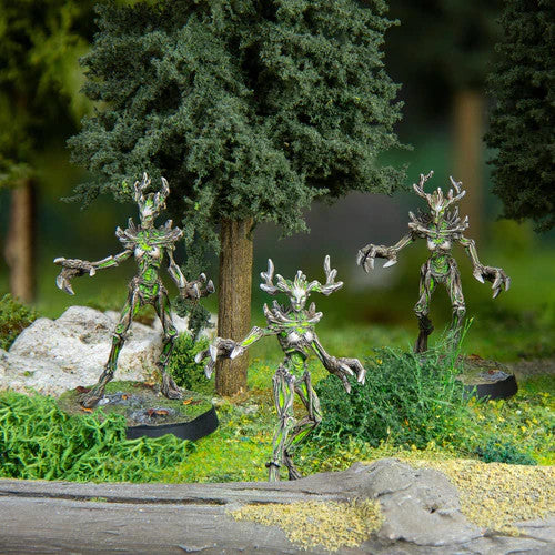 Three Modiphius The Elder Scrolls Call To Arms Spriggans miniatures, with branch limbs and antlers, stand among foliage in a forest diorama, their bark- and leaf-textured bodies evoking the games iconic creatures.
